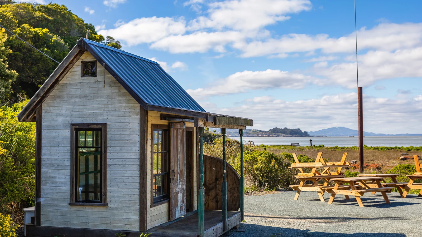 A small coastal timber cabin with a blue roof beside picnic tables, with the sea beyond.