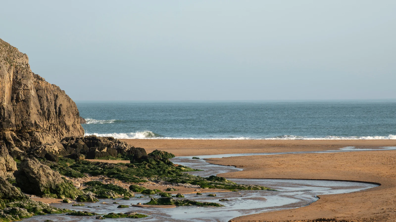 A sandy beach with rocky cliffs, shallow tidal channels and the sea beyond.