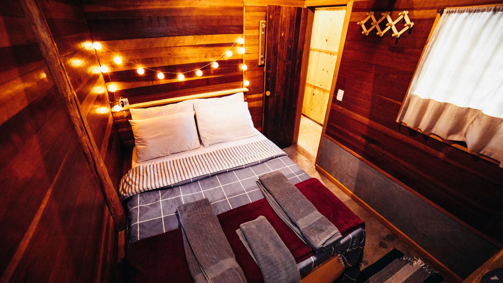 A compact timber-lined bedroom with a double bed, folded towels and warm string lights.