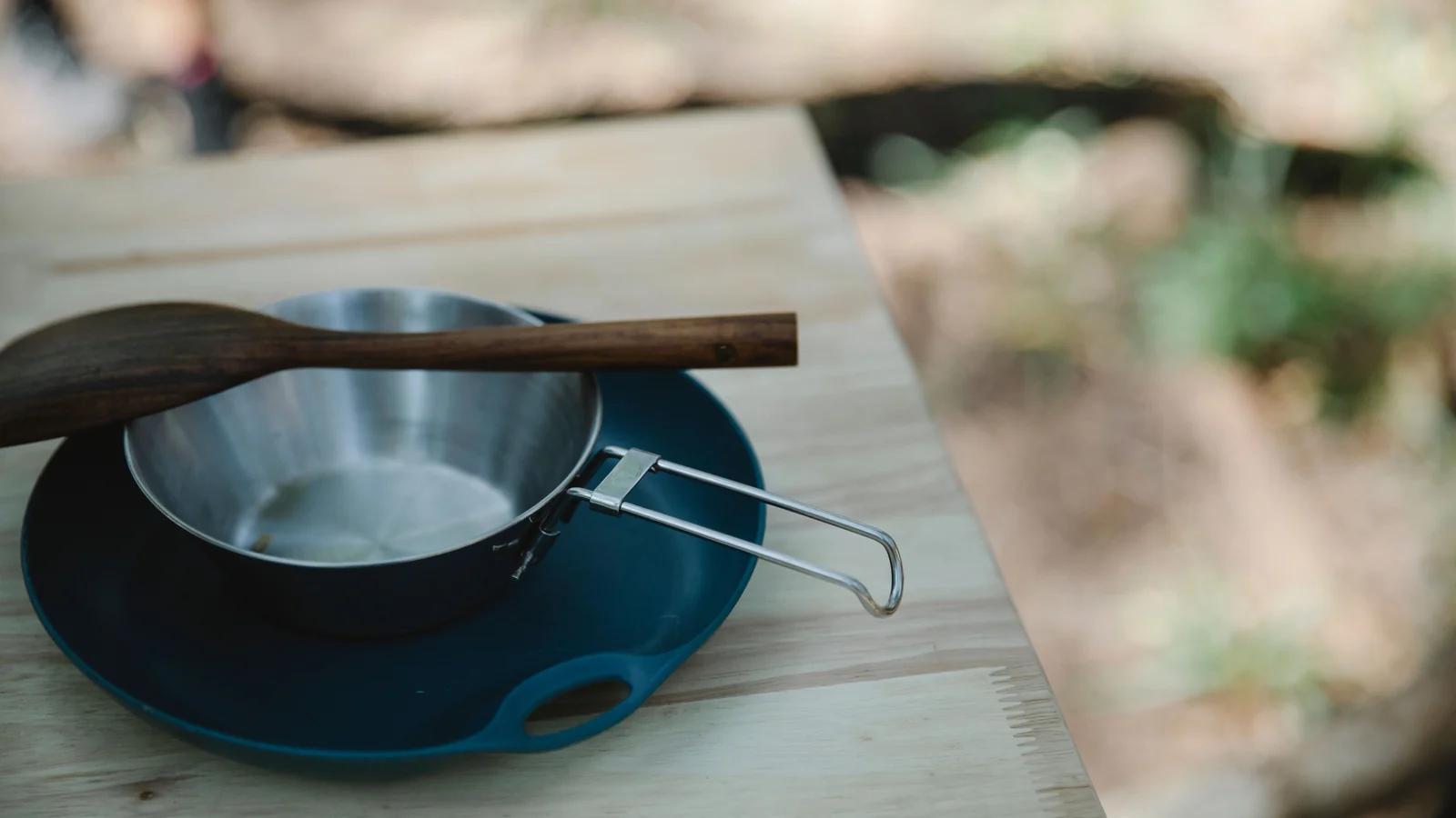 A metal camping pan and wooden spatula resting on a blue plate on an outdoor table.